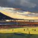 Painterly watercolour vista of a modern football stadium with players visible on the pitch under a dramatic warm sky, English Women Super League closing weekend atmosphere